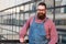 Bearded stylish craftsman wearing blue overalls looking at camera, outdoors. Family workshop, old manufactory worker