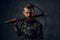 A bearded tattooed hipster guy in military shirt posing with a medieval crossbow. Studio photo against dark wall