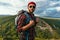 A bearded traveler with a backpack on the top of a mountain. Portrait of a traveler in a red cap and sunglasses.