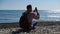 bearded white male photographer sits on stone beach in pink sweater with backpack over his shoulder and photographs