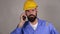 Bearded worker in yellow helmet talking on the phone with someone on grey background