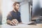 Bearded young coworker working at sunny office. Man using contemporary desktop computer and making notes in notebook