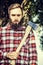 Bearded young man in checkered shirt holding a old ax on tree nature background, outdoor
