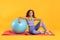 Beautiful African American woman with headphones and sports equipments sitting on yoga mat against yellow background