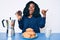 Beautiful african woman eating breakfast holding cholate donut pointing thumb up to the side smiling happy with open mouth