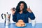 Beautiful african woman eating breakfast holding cholate donut smiling happy and positive, thumb up doing excellent and approval