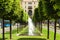 Beautiful alley with fountains. Summer hot day at city. Green trees and green grass among traffic street. Small fountains