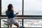 Beautiful alone brunette woman in a denim jacket and shorts sits on a high chair and looks out the huge window of her apartment.