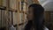 Beautiful Asian biologist student is walking along bookshelves in library indoors.