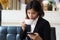 A beautiful asian business woman sitting on sofa drinking cup of hot coffee and using smart phone.