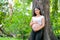 Beautiful Asian pregnant woman smile, lean at the tree and touch her belly with look forward among green garden with morning light