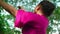 A beautiful Asian woman does a dancing ritual while wearing a pink dress and makeup in front of the big tree full of green bushes