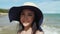 Beautiful Asian woman wearing a wide-brimmed hat, smiling, playing for the camera on the beach.