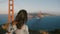 Beautiful back view of young tourist woman with backpack, hair blowing in the wind enjoying sunset at Golden Gate Bridge