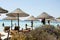 Beautiful beach resort with straw umbrellas on a blue sky and white clouds. On the background some people having fun on the beach