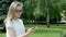 Beautiful bespectacled young woman smiles merrilly typing on her smartphone standing outdoors in a park in front of lawn