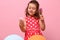 Beautiful Birthday girl in stylish pink dress with polka dots, combs her curly hair, admires her reflection on small pink cosmetic