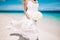 Beautiful blond bride in white wedding dress with white wedding bouquet stand on sea shore. Tropical turquois sea on the backgroun