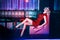 Beautiful blond model in red short fitted sequin dress relaxing on the square sofa in night club