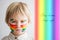 Beautiful blond toddler boy with rainbow painted on his face and messy hands