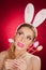 Beautiful blond woman as Easter bunny with rabbit ears on red background, studio shot. Young lady holding three colored eggs