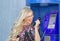 A beautiful blond woman wiaring glasses is holding a telephone receiver in a payphone. Emotionally shouts into the phone.
