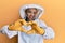 Beautiful blonde caucasian woman wearing protective beekeeper uniform smiling in love doing heart symbol shape with hands