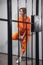 A beautiful blonde criminal in an orange robe stands in her cell behind bars, leaning against the wall