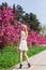 Beautiful blonde girl with long wavy curls in a white dress walk in the park among the pink trees, stunning makeup