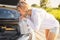 Beautiful blonde girl with oil canister is perplexed looking at the car engine on a country road in the rays of the setting sun