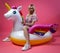 A beautiful blonde girl in a sexy sundress with slim legs in white sneakers sits on an inflatable multi-colored unicorn on a pink