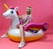 A beautiful blonde girl in a sexy sundress with slim legs in white sneakers sits on an inflatable multi-colored unicorn on a pink