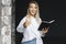 Beautiful blonde girl is standing in white shirt on dark background near brick wall in office, shows approving gesture.