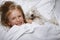 Beautiful blonde little girl laughing and lying with white schnauzer puppy dog on white bed. Friendship concept.