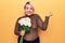 Beautiful blonde plus size woman holding bouquet of flowers over isolated yellow background screaming proud, celebrating victory