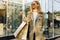 Beautiful blonde in sun goggles and shopping bags stands on against a glass building, in a beige raincoat