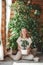 A beautiful blonde woman in beige clothes cares for a ficus flower against the background of a large number of house