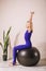 A beautiful blonde woman in a closed sports jumpsuit of blue color, practicing yoga, sits on a fitball with her arms raised