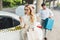 beautiful blonde woman in eyeglasses using smartphone while man putting suitcase in trunk of taxi car