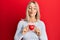 Beautiful blonde woman holding heart making fish face with mouth and squinting eyes, crazy and comical