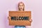 Beautiful blonde woman holding welcome doormat in shock face, looking skeptical and sarcastic, surprised with open mouth