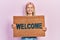 Beautiful blonde woman holding welcome doormat smiling with a happy and cool smile on face