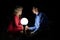 Beautiful blonde woman in red dress, and a man in blue shirt sits together in darkness, next to a ball shaped lamp. Couple dating