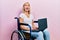 Beautiful blonde woman sitting on wheelchair holding laptop angry and mad screaming frustrated and furious, shouting with anger