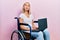 Beautiful blonde woman sitting on wheelchair holding laptop looking at the camera blowing a kiss being lovely and sexy