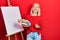 Beautiful blonde woman standing drawing with palette by painter easel stand smiling and laughing hard out loud because funny crazy