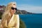 Beautiful blonde woman, standing in an elegant jacket and sunglasses by the sea