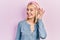 Beautiful blonde woman standing over pink background smiling with hand over ear listening an hearing to rumor or gossip