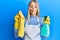 Beautiful blonde woman wearing cleaner apron holding cleaning products making fish face with mouth and squinting eyes, crazy and