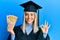 Beautiful blonde woman wearing graduation cap and ceremony robe holding philippine pesos doing ok sign with fingers, smiling